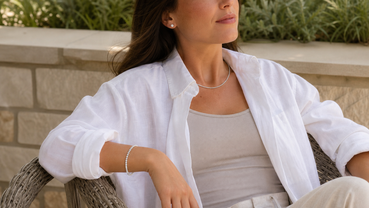 Woman in a white shirt wearing silver tone stud earrings, a cable chain necklace, and a cubic zirconia tennis bracelet outdoors.