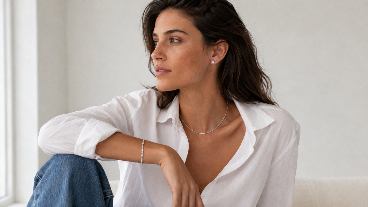 Woman in a white shirt and blue jeans wearing round clear cubic zirconia stud earrings, a narrow silver-tone tennis bracelet, and a fine silver-tone cable chain.
