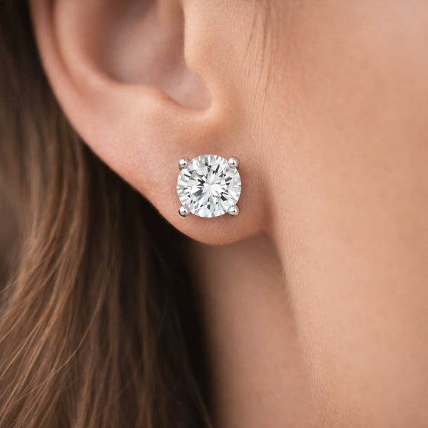 Worn view of a round cubic zirconia stud earring in silver tone shown on the ear highlighting the faceted stone brilliance and polished four prong setting.