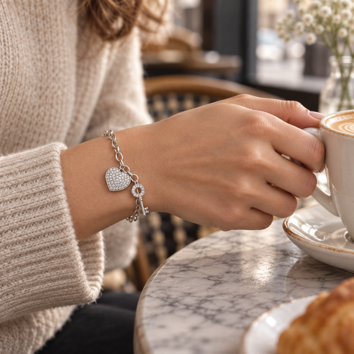 Woman reaching for a coffee cup while wearing a silver-tone heart and key charm bracelet.