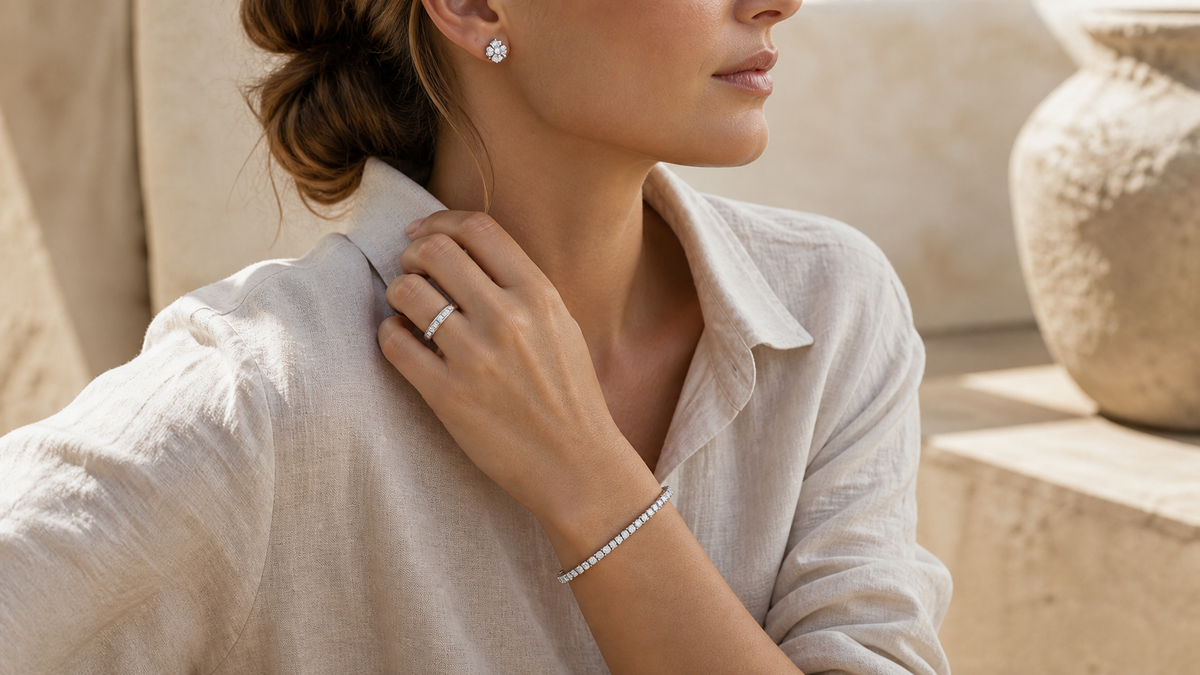 Woman in a pale linen shirt wearing round silver-tone cubic zirconia studs, a silver-tone tennis bracelet, and a rectangular-stone band ring.